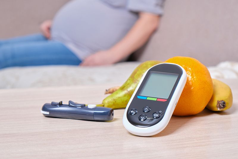 fruit and glucose meter on table, pregnant woman on sofa, blood sugar control concept
